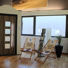 Moderne Ausstellung mit Holztür, Liegestühlen und großen Fenstern in hellem Showroom.